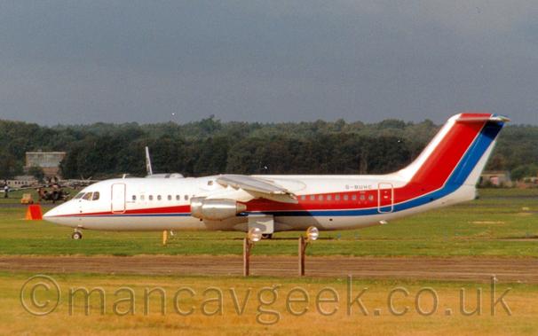 Side view of a high-winged, 4 engined jet airliner taxiing from right to left with flaps drooping behind the wings, suggesting it has just landed.
The plane is mostly white, with  a thin red and blue stripe running along the body, getting wider as it moves rearwards, before sweeping up to fill the tail.
The blue registration "G-BUHC" .
Yellowing grass fills the foreground at the bottom of the frame, with what appear to be lights on a pair of brown wooden poles next to a taxiway.
Several other planes can be seen in the background behind this planes nose, with rows of trees in the distance under grey-blue sky.