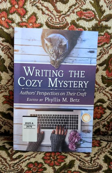 Phyllis M. Betz' essay collection "Writing the Cozy Mystery" propped up on a cozy chair with a vintage orange and green flower pattern (very cozy!)