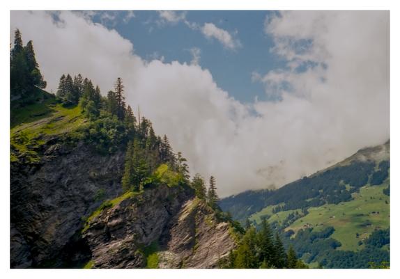 Colour photograph of a steep rocky cliff covered with green pines in the Swiss Alps, with rolling green hills below and thick white clouds drifting through the valley under a bright blue sky.
