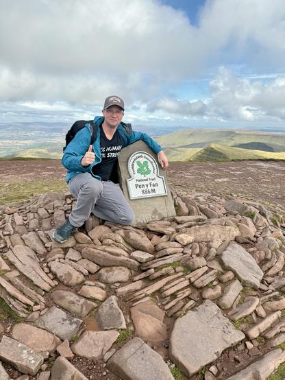 It’s me. In a grey cap, blue coat, t-shirt with amusing “3% human 97% stress” written on it. At the top of Pen Y Fan. Behind me is the Bannau Brycheiniog National Park.