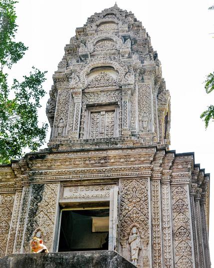 Photo shows a temple tower with an intricate and detailed outer decoration. Some visible carvings are of standing figures surrounded by flowers and lotus bulbs. Over the main room with an open entrance door, the top of the temple has a closed small gate echoing the main entrance. Then it continues in late Bayon style. Then it continues in seven decreasing size levels to the top spire. In front there are stairs divided in 2 directions. Fronted by a large cement block, a slightly curious looking male Macaque monkey is halfway hidden. Cambodia is as safe and kind as ever. Angkor and Siem Reap welcomes visitors. Enjoy the Kingdom of Wonder.