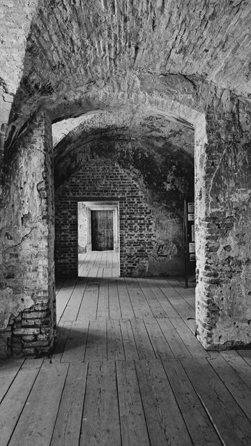 A black and white photo shows a long, empty room with a wooden floor. The room is oriented horizontally, with the point of view from the middle of the room. The ceiling is made of rounded brick arches. Two large, weathered brick pillars stand on either side of the room, near the front. There is a doorway in the far wall, framed by brick, which leads into a similar empty room with a closed wooden door.
