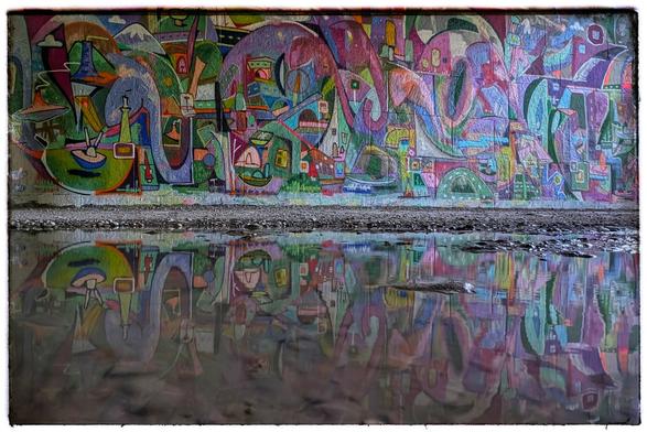 Eine abstrakte, bunte Wandmalerei, die die ganze Wand ausfüllt und sich in einer Pfütze spiegelt.