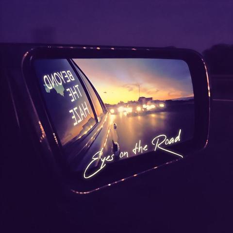 A photo of a car's side mirror, taken from inside the car during dusk. The mirror shows several cars driving on a freeway, behind them a sunset, and also a part of the car the mirror is attached to. The artist name "Beyond the Haze" is edited into the car window, mirrored. At the bottom of the mirror it says "Eyes on the Road" in a handwritten font.