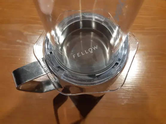 The image shows an AeroPress coffee brewer positioned on top of a metal pitcher with a handle. Inside the AeroPress, there is a Fellow metal filter. The AeroPress is transparent, allowing a view of the filter and the word "FELLOW" engraved on the bottom. The surface beneath is wooden, and the overall setup appears clean and organized.