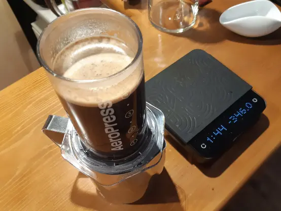 The image shows an AeroPress brewer positioned on top of a metal pitcher with a handle. The AeroPress is filled with brewing coffee, and its top is open, revealing the coffee inside. Next to the AeroPress, there is a digital scale. The background features a wooden surface, and there are additional items, including a glass container and a small white dish, partially visible.