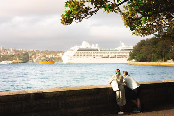 Two people lean against a stone wall overlooking a body of water. A large white cruise ship sails in the background. A tree with green leaves frames the top right corner. The opposite shore features a distant cityscape under a cloudy sky.