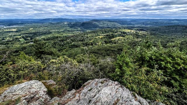 We are on a mountain standing atop a rock outcrop of light gray rock marbled with darker gray. We are looking out over the tops of some pine trees and hardwoods. We are looking across a broad valley hundreds of feet below filled with the greens of late summer. Most of it is covered with deep green forests broken by emerald green pastures and farm fields. A pair of almost adjacent ponds are to the left of our view. A line of blue green hills and mountains runs along the far horizon. A few smaller wooded hills are scattered over the valley floor. A morning of clouds is just giving way to bright sunshine. The sky is a soft blue filled with lines of retreating clouds.