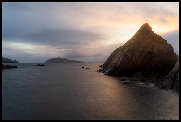 Long exposure sunset photograph of dramatic rocky coastline at Dunquin Harbour with large triangular rock formation in foreground and layered mountain silhouettes across the water under moody evening sky, County Kerry.