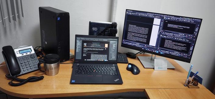 En el centro de un escritorio de madera clara, se encuentra una laptop Lenovo ThinkPad con la pantalla encendida, mostrando un entorno de escritorio Fedora KDE Plasma. El tema oscuro "Breeze Dark" se destaca en la pantalla de la laptop, que parece estar ejecutando un navegador web con información técnica.

A la derecha, un monitor externo de mayor tamaño amplía el espacio de trabajo. También con el tema oscuro activado, la pantalla del monitor muestra una ventana dividida en varias secciones, lo que sugiere una tarea de escritura o edición con múltiples documentos o paneles abiertos simultáneamente. Debajo del monitor, se encuentra un ratón ergonómico, un pequeño teclado y un calendario.

Junto a la laptop, se aprecia una torre de PC color negro. El escritorio también está equipado con herramientas de comunicación y trabajo, incluyendo un teléfono de oficina, una taza de café metálica, y un portalápices. En el fondo, una persiana de color claro contrasta con los tonos oscuros de los equipos.