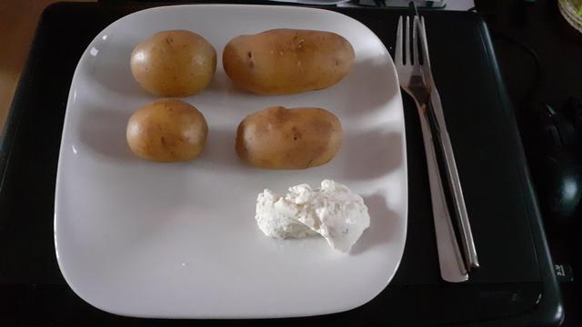 Ein weißer quadratischer Keramikteller mit 4 Pellkartoffeln und etwas Quark auf dem Teller, rechts daneben liegt das Besteck. Alles steht und liegt auf einem zu geklappten Notebook auf einem dunkelbraunen Couchtisch.