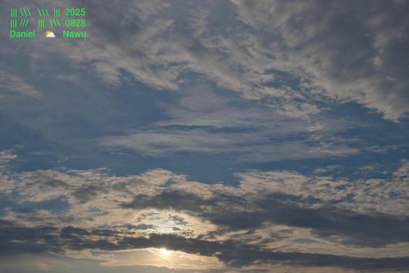 Fotografie des Himmels im Querformat. Das obere und untere Drittel des Fotos sind von unterschiedlich aussehenden Wolken ausgefüllt. Mittig im unteren Drittel scheint die Sonne durch diese Wolken. Das mittlere Drittel des Bildes ist vorwiegend blauer Himmel, in dessen Zentrum sich aber ganz ungewöhnlich aussehende, sehr zarte Wolken befinden. Anders als die anderen Wolken sind sie durch die Lichtbrechung in lila, grüne, blaue und ein wenig gelbe Töne getaucht. Das erinnert ein bisschen an Perlmutt. Daher auch der Name Perlmuttwolken.

Oben links in der Ecke befindet sich mein giftgrünes Wasserzeichen.