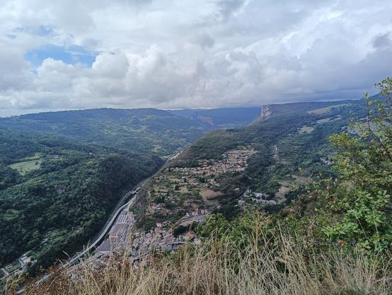 Vue panoramique prise en haut d'une falaise, avec une vue dégagée sur une vallée au fond de laquelle il y a un bourg avec des bâtiments industriels, des collines boisées et une autre falaise abrupte. Le ciel est bleu avec de nombreux nuages blancs.