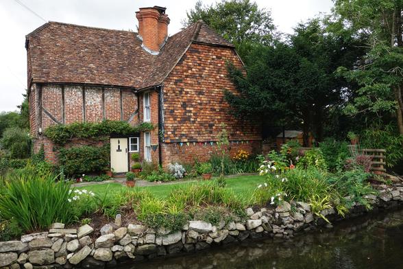 A quaint, historic cottage made of red brick and timber-framing, with a gabled, tiled roof. The house stands beside a calm river or stream, its banks fortified by a low wall of grey stones. The front of the cottage is decorated with a small yellow door, and a string of colourful bunting hangs across the front lawn. The garden is lush and vibrant, with a mix of green shrubs and bright flowers in various colours, including white, yellow, and orange. The scene is framed by a variety of mature trees and foliage.