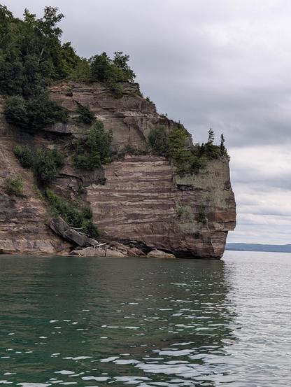 A picture of a rock feature on the western side of Grand Island. It's part of the Pictured Rocks Lakeshore. The cliffs resemble a helmet of some sort facing to the right stop the water. It's not dissimilar to an Amtrak cabbage car. The rock face itself is striped muted burgundy and muted sandy yellow/cream. Various plants and trees cover the top of most of the rocks, including a sharp thin slope down one side of the helmet feature, nearly reaching the cold teal green water. Even with a disrupted surface, the downward visibility is excellent and you can see the shallow floor in this section. There is a steep drop off as you go farther from shore.