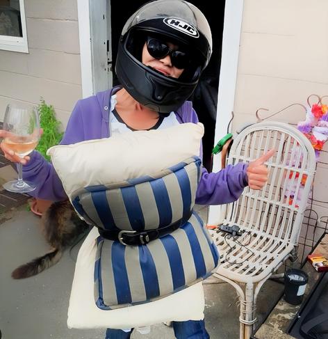 Woman wearing a motorcycle helmet and sunglasses, posing with a pillow around her waist belted with a black belt, holding a glass of wine in one hand and giving a thumbs up with the other, standing in front of a door with a white wicker chair nearby.