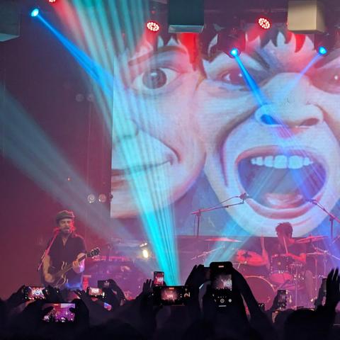 Una vista frontal de un concierto de música en vivo. En el fondo, una pantalla gigante muestra una ilustración vibrante de dos rostros humanos caricaturescos y exagerados: uno sonriente con la boca cerrada y el otro con la boca abierta en un grito o canto, con dientes blancos visibles. Rayos de luz azul brillante y rojo intenso cruzan el escenario y la pantalla, creando una atmósfera dinámica. En el centro-izquierda del escenario, un guitarrista con barba y un sombrero oscuro (posiblemente un fedora o boina) toca una guitarra eléctrica. Parcialmente visible a la derecha, un baterista toca la batería. En primer plano, desde la perspectiva de la audiencia, se ven numerosas manos levantadas sosteniendo teléfonos móviles, grabando o fotografiando el espectáculo.