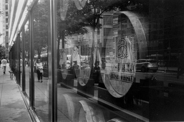 Black and white photo of a floor to ceiling window panes from the side walk level. The window is reflecting the outside world, prominently a person walking towards the camera in the left third of the frame. The rest of the frame is looking through the window unto wooden barrels on their side, the flat bottom of just one of the barrels is legible "UISCE BEATHA" (which is what whiskey is called in Irish and it means "water of life") on top and "IRISH WHISKEY" on the bottom.