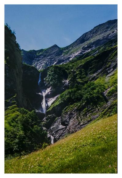 Color photograph from the Swiss Alps. It shows a waterfall photographed from Tschinglenalp. In the foreground is a steeply rising green meadow, and next to and behind the waterfall are partly wooded rocky cliffs.