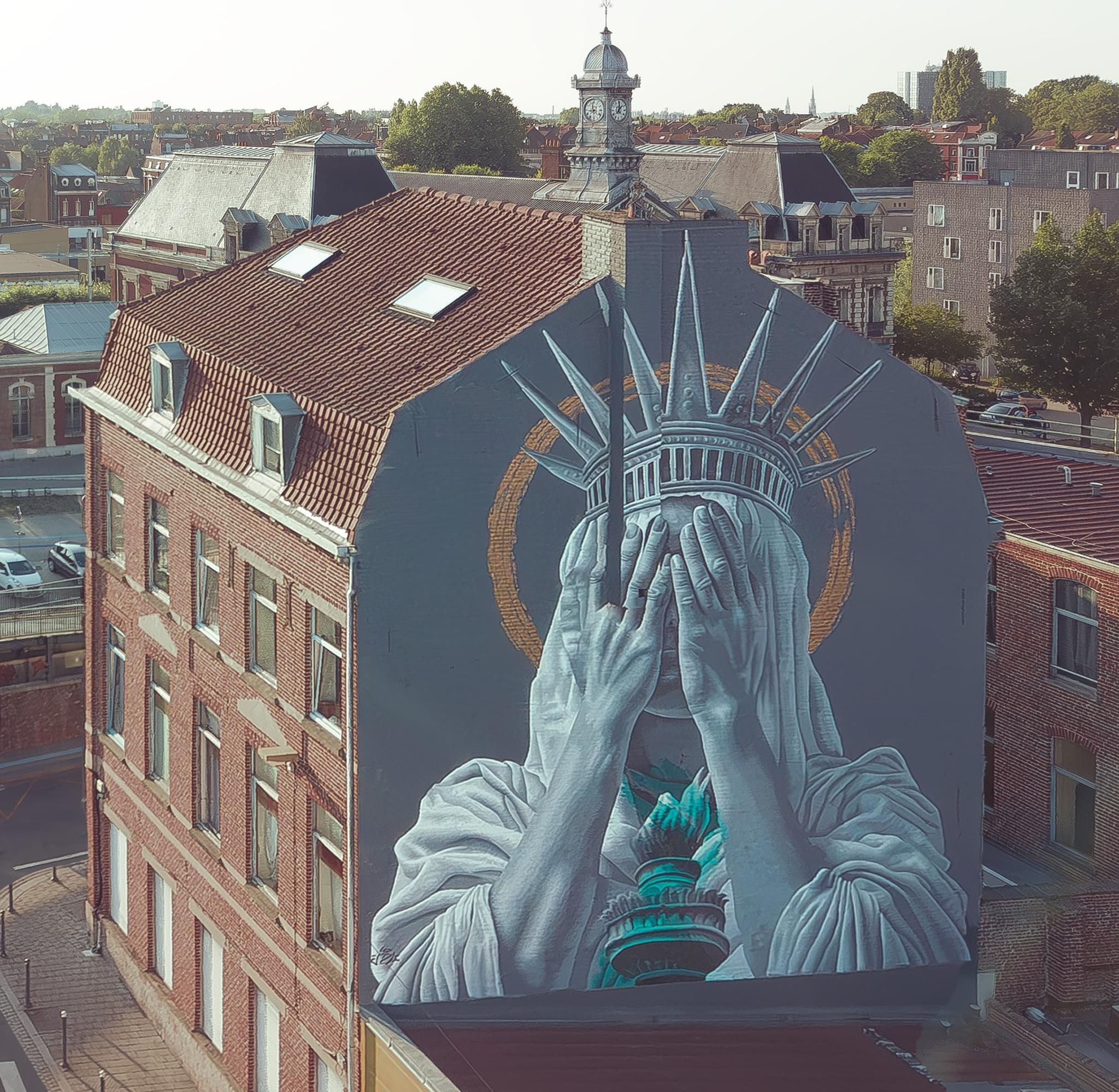 Mural by JDL Street Art in Roubaix, Lille, France, depicting the Statue of Liberty covering her face with both hands in a gesture of shame. The figure is painted in shades of grey with teal accents on the torch and crown, set against a muted background with a golden halo-like circle. The artwork spans the side of a tall brick building, contrasting modern street art with historic European architecture in the surrounding cityscape.