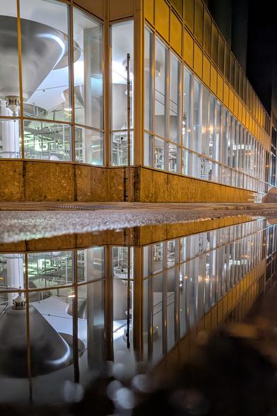 Das Bild zeigt ein modernes Industriegebäude bei Nacht. Die Architektur ist geprägt von großen Fenstern, durch die das hell erleuchtete Innere sichtbar wird. Im Inneren befinden sich zahlreiche metallische Maschinen und technische Anlagen, die ordentlich angeordnet sind und einen funktionalen Eindruck vermitteln. Die Umgebung außerhalb des Gebäudes ist dunkel, wodurch die Beleuchtung im Inneren besonders hervorsticht. Im Vordergrund des Bildes liegt eine Pfütze auf dem Boden, die das Gebäude und dessen beleuchtetes Inneres klar und symmetrisch widerspiegelt.