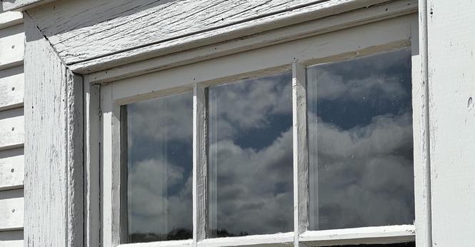 The top part of a mulitipane window reflects a blue sky filled with white cumulus clouds. The window frame and siding is white wood with very prominent wood grain patterns, and the lighting emphasizes shadows to give shades of grey. [Signal Hill National Historic Site (St. John’s, Canada) August 2025]