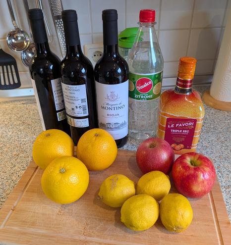 Auf einer Küchenanrichte liegt ein Brett mit einigen Orangen, Zitronen und Äpfel. Dahinter stehen ein paar Flaschen Rotwein, ein Likör und Limonade.
