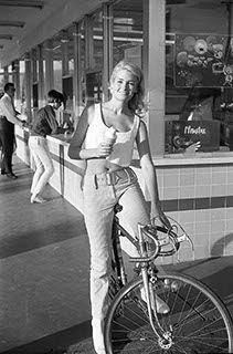 Black-and-white photo of a young Deidre Hall from the 1960s, standing with a bicycle in front of a diner counter, wearing a sleeveless top and high-waisted pants, holding an ice cream cone, capturing a casual and stylish moment from her early modeling portfolio.