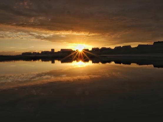 Gegenlicht Aufnahme mit Sonnenstern von häusershilouette im orangen Abendhimmel, die sich im See spiegelt
