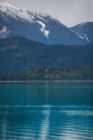 A vertical landscape. A tiny white dot in the center - the lighthouse - is surrounded by vast mountains, some snow-capped. That tableau is reflected in the rippling turquoise waters of Kootenay Lake.