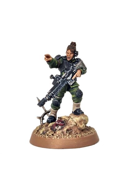 Photograph of a Minka Lesk Imperial Guard miniature from Games Workshop on a white background. The model wears the usual Cadian style armour, painted black with green fatigued and light brown gaiters. She is pointing toward the camera, holding her gun in a ready position.