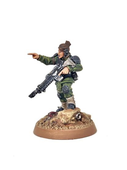 Photograph of a Minka Lesk Imperial Guard miniature from Games Workshop on a white background. The model wears the usual Cadian style armour, painted black with green fatigued and light brown gaiters. She is pointing with two fingers, holding her gun in a ready position.