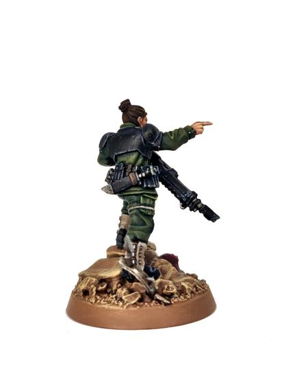 Photograph of the back of a Minka Lesk Imperial Guard miniature from Games Workshop on a white background. The model wears the usual Cadian style armour, painted black with green fatigued and light brown gaiters. She is pointing toward the camera, holding her gun in a ready position.