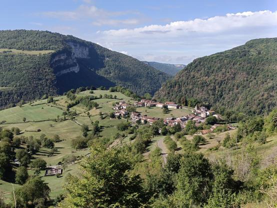 Vue panoramique avec en contrebas un village circulaire (Oncieu) dont le centre est une prairie, et un petit cimetière carré. De part et d'autre, des prairies vallonnées, des reliefs escarpés et boisés, avec des falaises à gauche. Le ciel est bleu avec des nuages blancs.