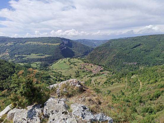 Vue panoramique similaire à la précédente, mais prise de beaucoup plus haut à la croix d'Évosges. Au premier plan, des rochers et au loin, le village circulaire d'Oncieu. Le ciel est bleu avec de plus en plus de nuages blancs et gris.