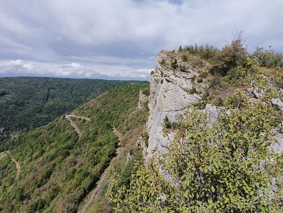 Vue panoramique prise des rochers de la croix d'Évosges, avec un des rochers proches et sa falaise calcaire, et en contrebas les lacets du chemin emprunté pour venir, entre vignes et bois. Le ciel est couvert mais encore lumineux.