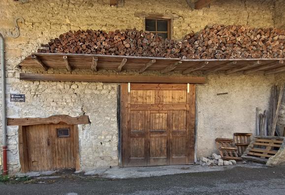 Vue rapprochée d'une grange en pierre claire, avec à gauche une petite porte en bois surmontée d'une poutre tordue, au centre une porte de grange en bois décrite dans le pouet, et par dessus une galerie en bois (qui s'appelle un dréfiat) où sèche du bois.