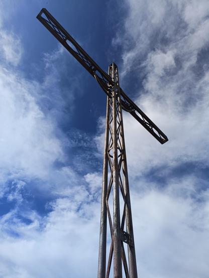 Vue en contre-plongée d'une grande croix en métal avec des montants en croisillons. Le ciel est bleu avec des nuages blancs.