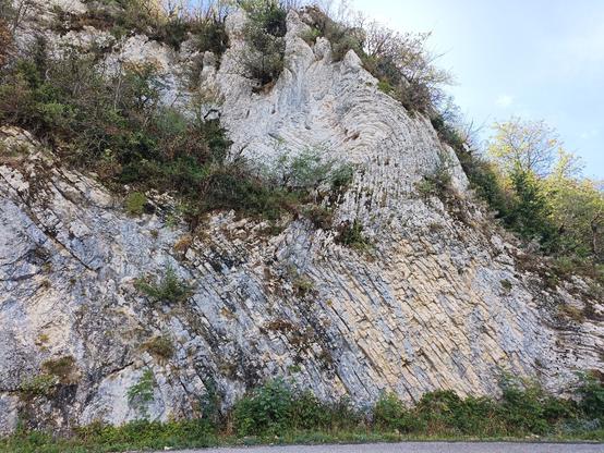 Vue rapprochée d'une paroi rocheuse de couleur claire, dont les couches sont quasiment à la verticale avec un plissement d'une partie d'entre elles. Le ciel est bleu avec une lumière un peu à contre-jour.