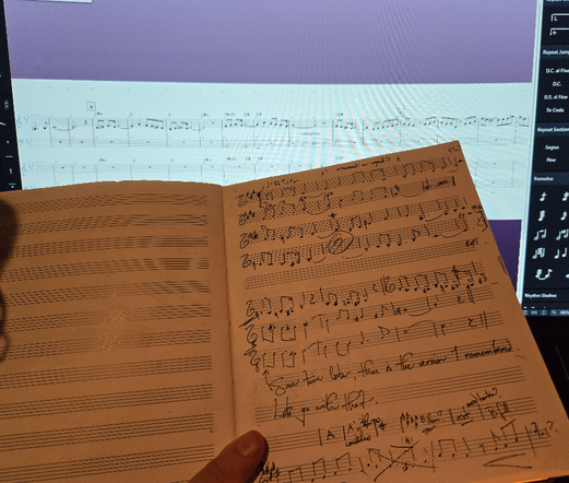 Holding a music notebook with handwritten notes in front of a computer monitor with music software open. Emmanuel is transcribing the handwritten music into the computer for later stages of production.
