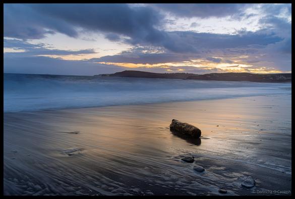 Long exposure photograph of a driftwood log on wet sand at Owenahincha Beach with smooth water and dramatic cloudy sky after sunset, showing golden reflections on the beach surface, County Cork.