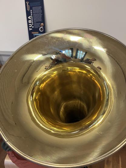 An overhead view of a shiny brass tuba bell, reflecting the surrounding room and people. In the background, a sign with information about tubas is visible.