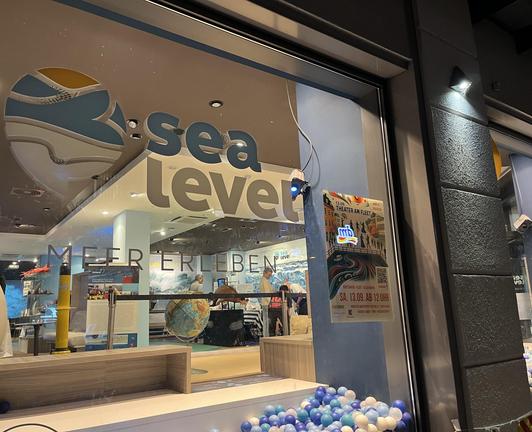 A glass storefront featuring the logo "sea level" along with the phrase "MEER ERLEBEN." The interior displays ocean-themed decorations, including a globe, and there is an event poster for "Theater am Fleet" nearby. The scene is