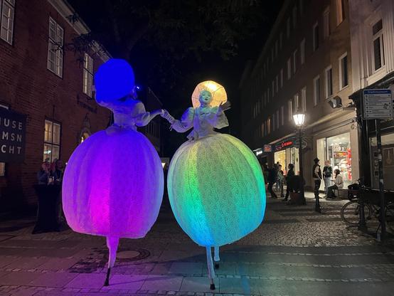The image shows two illuminated performers on stilts, dressed in elaborate outfits with large, colorful skirts. They are standing in an urban setting at night, with shops and a crowd visible in the background, creating a festive atmosphere.