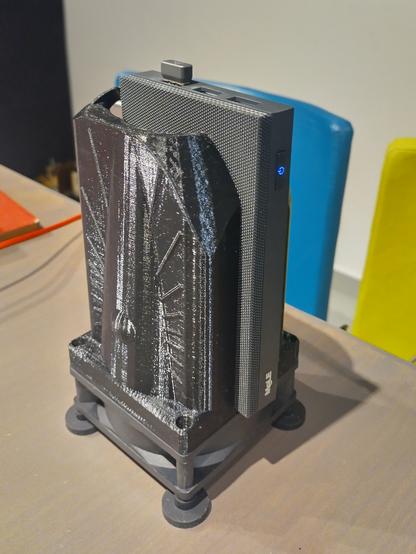 A 80 mm Computer fan with short rubber feet flat on a table. On top of it mounted is a Art Deco inspired tower like 3D-printed contraption. It acts as a fan duct encompassing a small flat micro PC that does not have its own cooling fans.