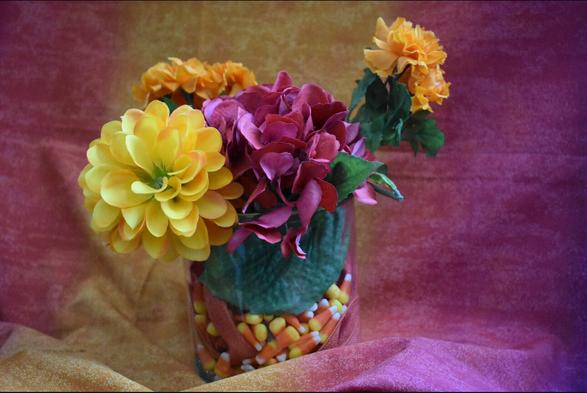 A handmade flower arrangement in a clear vase filled with real candy corn and wrapped with a ribbon. The arrangement includes yellow, orange, and deep red artificial flowers. Behind it is a textured background in shades of orange and maroon, creating a warm autumn feel.