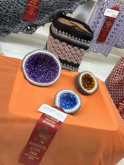 Crocheted geodes. Left is large and purple, middle is small and blue and far right is small and orange and they’re sitting on an orange table cloth.