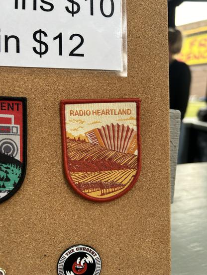 A red and yellow patch of a giant accordion looming in the background of rolling hills. It says Radio Heartland above it.