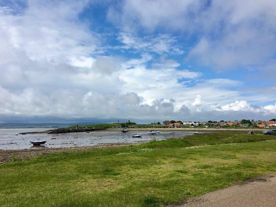 Along the Alternative Coast to Coast route near Holy Island in Berwick-upon-Tweed, a serene coastal landscape stretches beneath a dynamic sky of blue patches and billowing clouds. Small boats rest on the muddy shoreline, left idle by the retreating tide, their hulls contrasting against the dark, wet sands. A narrow pier juts modestly into the shallow water, adding a rustic charm to the scene. In the distance, a quaint village with red-roofed houses and white cottages nestles against the lush greenery, its tranquility mirrored by the calm waters. The expansive grassy foreground, dotted with wildflowers, merges seamlessly with the undulating coastline, while a few scattered cars hint at quiet human presence. The horizon is softened by misty hills, blending earth and sky in a peaceful, timeless panorama.