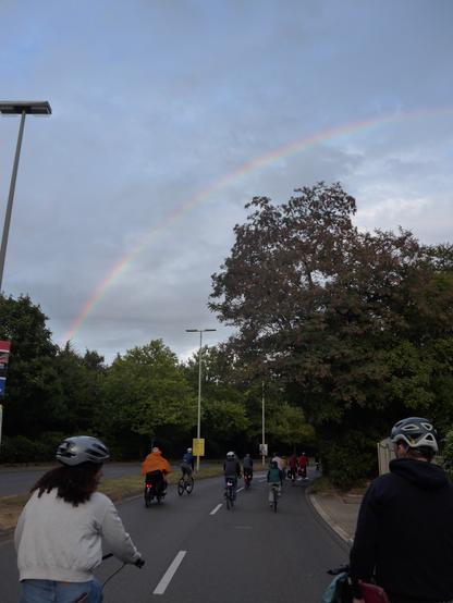 Menschen auf Fahrrädern fahren auf einer Straße, darüber ist ein Teil eines Regenbogens zu sehen